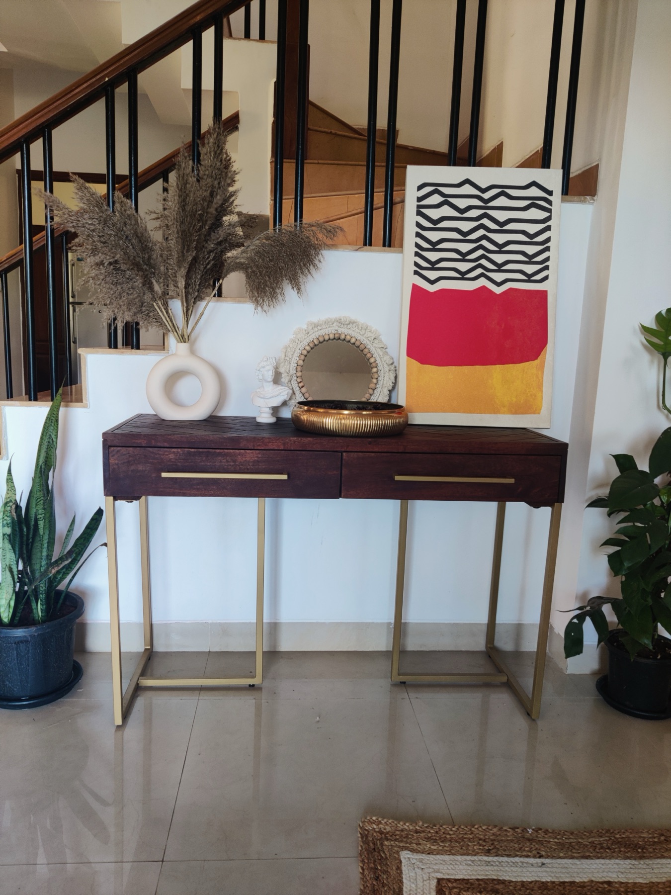 Burgundy console on brass legs below the stair, styled with pampas grass and a bold graphic canvas