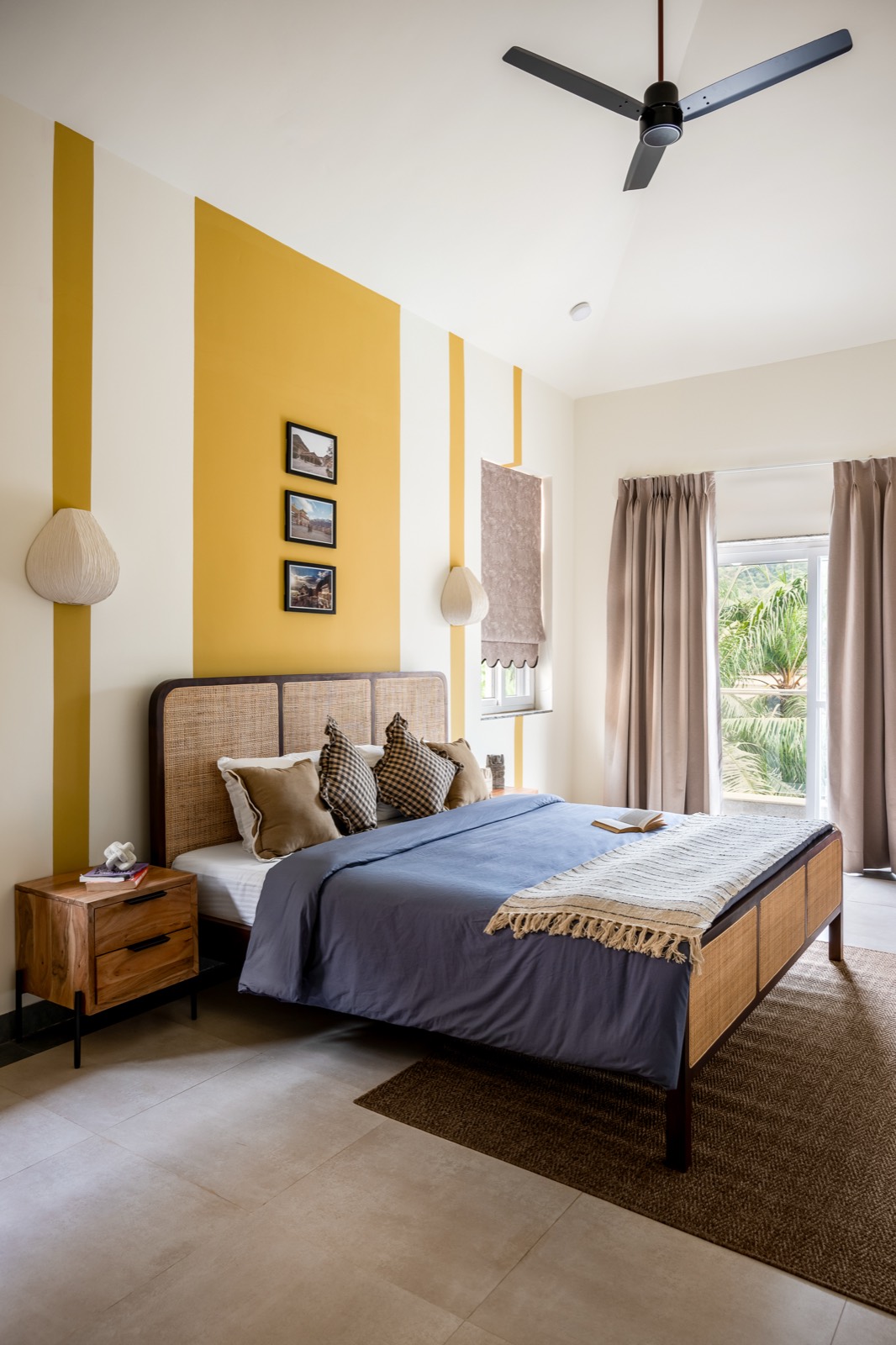 Bedroom with broad mustard and ivory bands behind a cane bed, woven wall lights and garden beyond