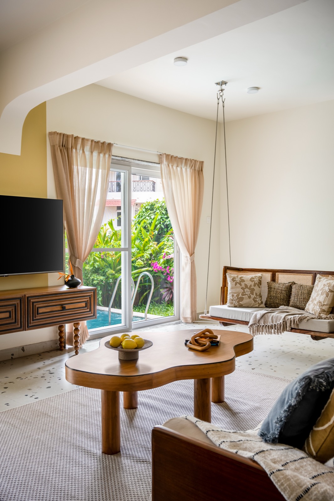 Living room opening to the pool and garden, with a cane swing, curved wooden coffee table and sheer curtains