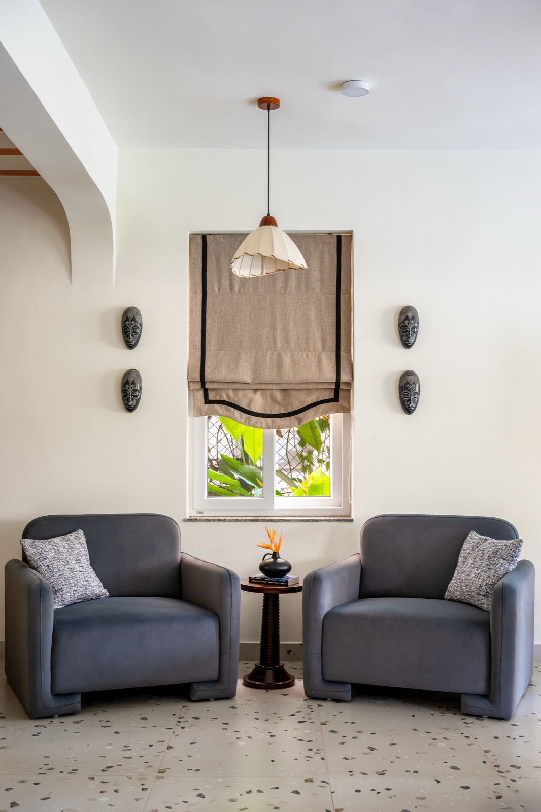 Two slate-grey armchairs flanking a small round table under a jute blind and carved wall masks