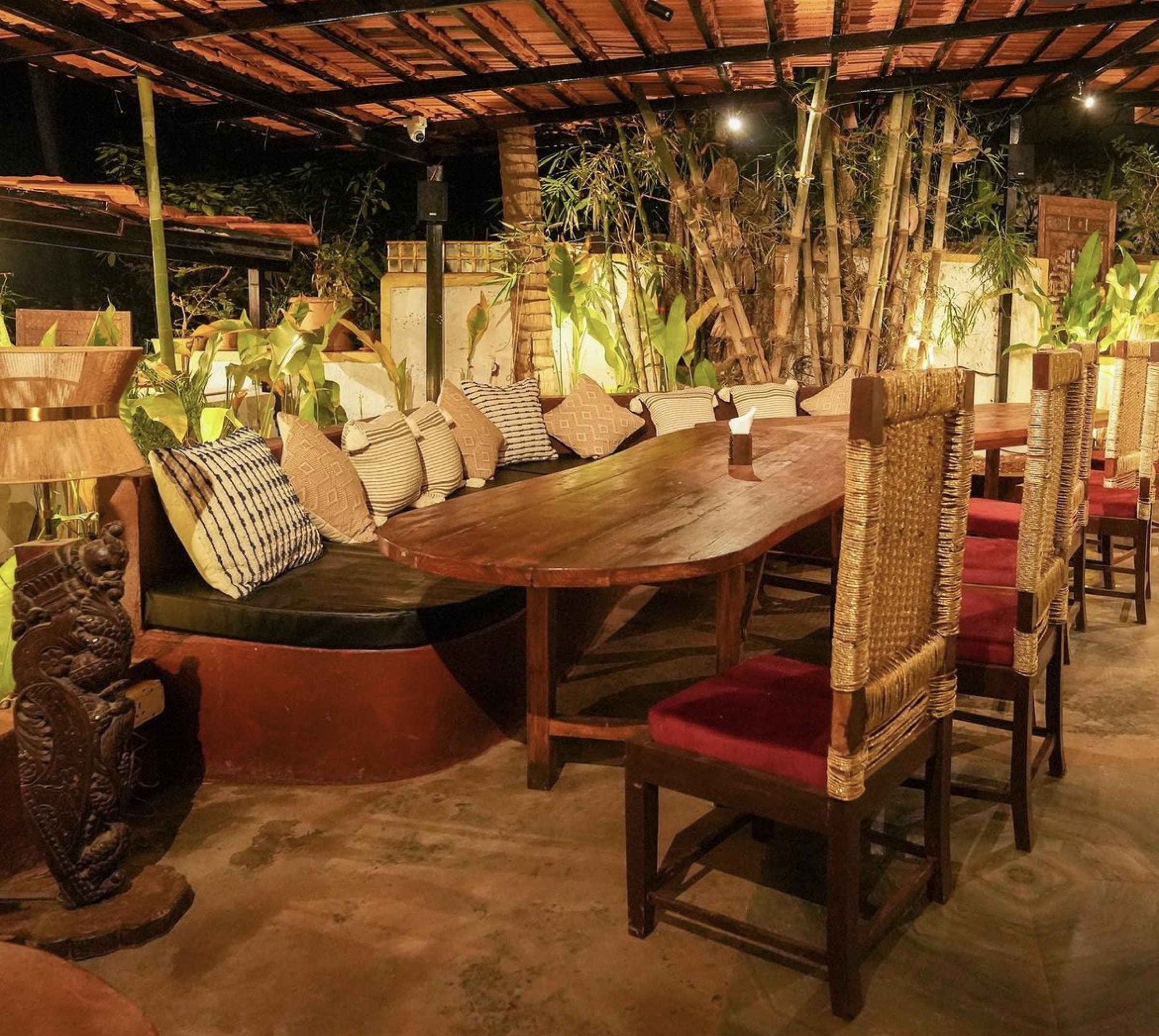 Long timber table with a cushioned bench and rush-backed chairs among bamboo