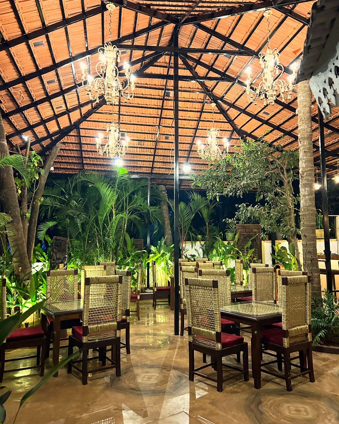 Dining tables and cane chairs under the clay-tiled roof, chandeliers and greenery overhead