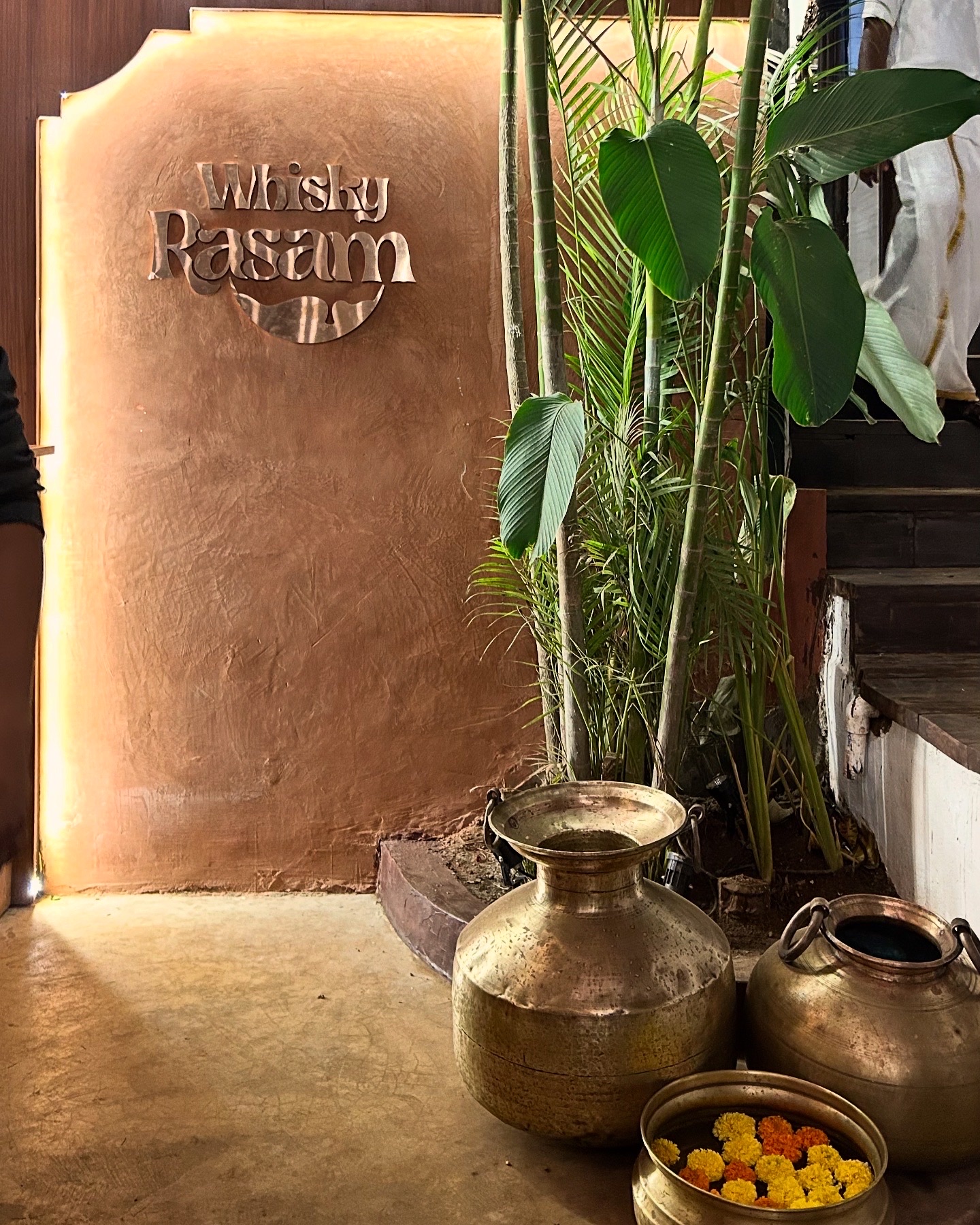 Whiskey Rasam signage raised on a rust-toned plaster wall above brass pots